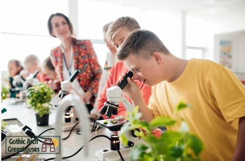 Students examining plant roots with a microscope inside a school greenhouse during a science lesson